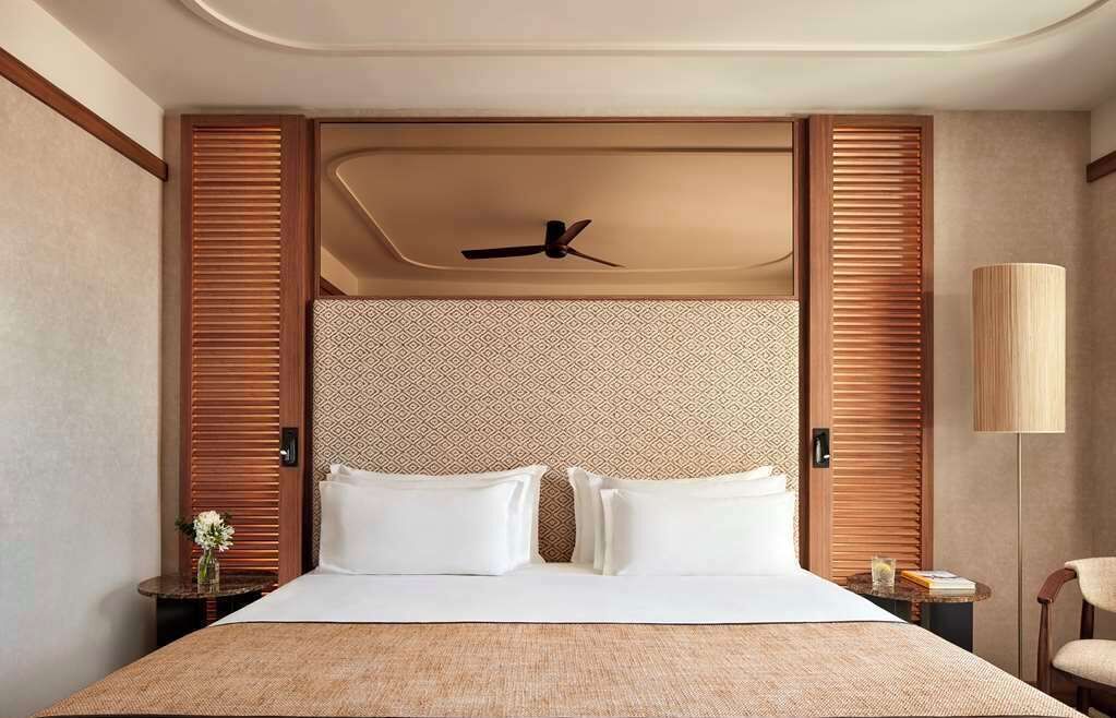 Camera d'albergo con un letto grande, una testiera in legno, una ventola a soffitto e un comodino con una lampada.