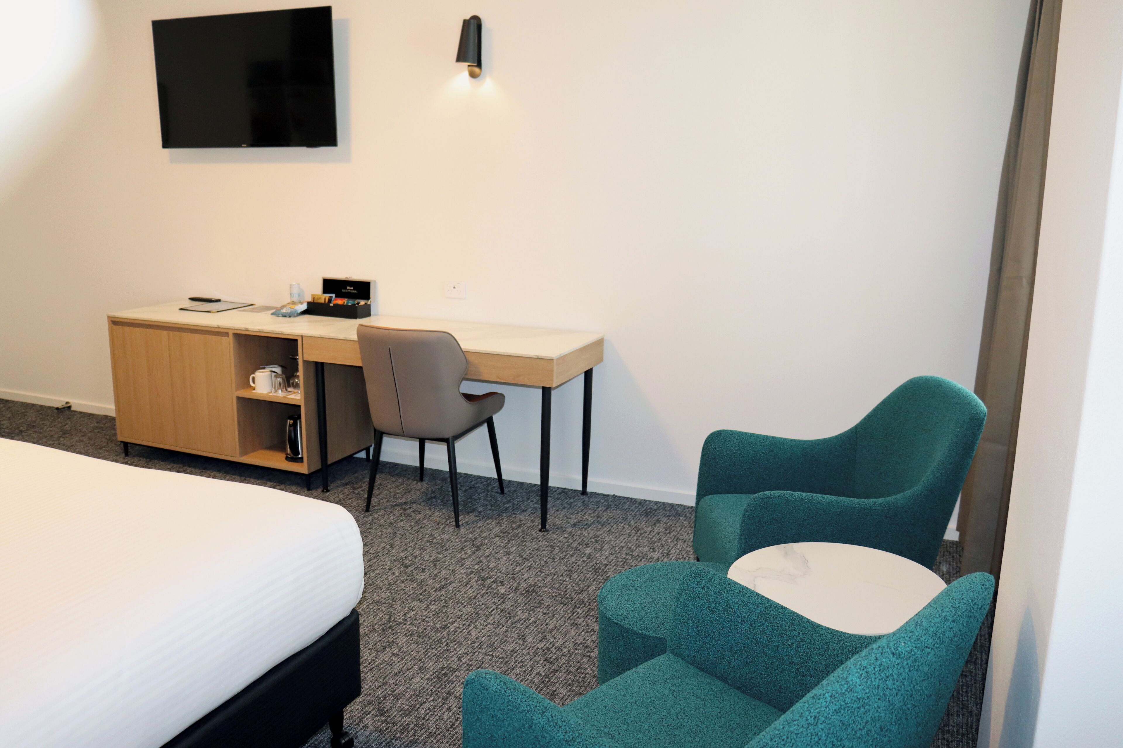 Habitación de hotel con cama, escritorio con silla, una mesita y televisor montado en la pared.