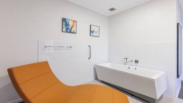A modern bathroom with a white bathtub, a curved orange chair, and two abstract paintings on the wall.