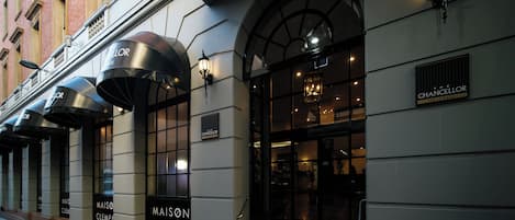 A hotel entrance with "Maison Clement" signage, large windows, and a covered entrance area.