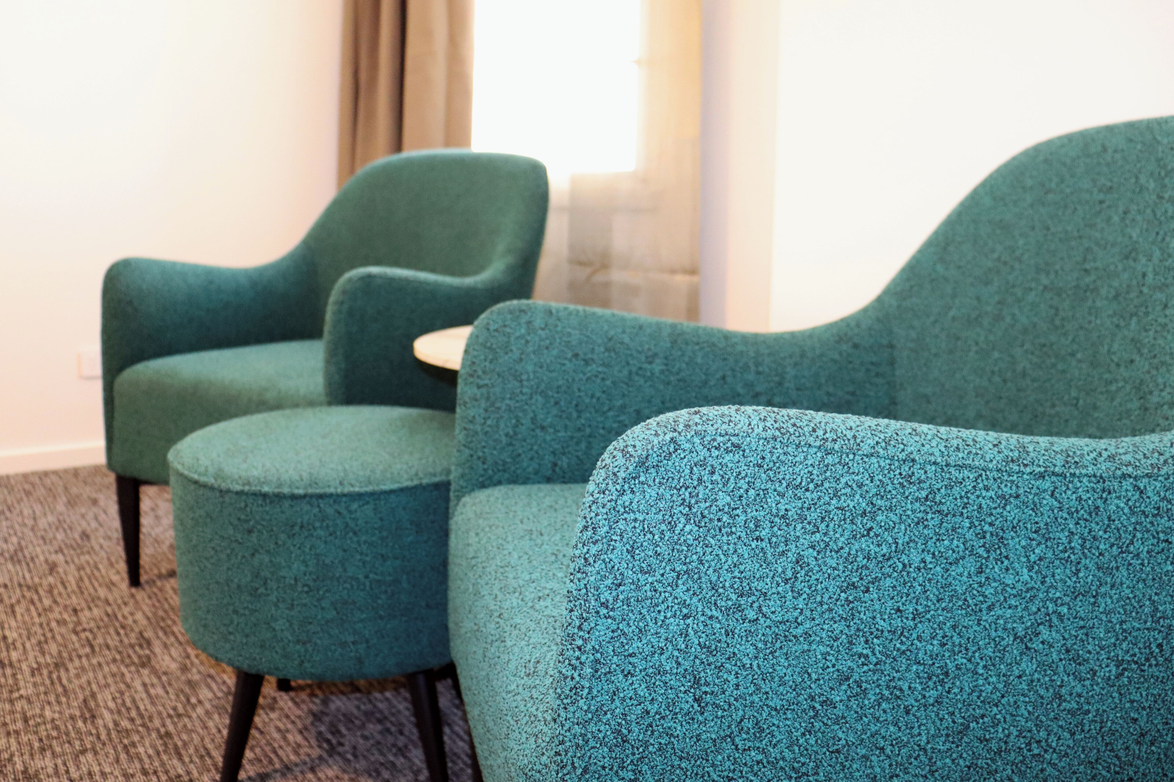 Dos sillones azul verdoso con patas de madera y una mesita, ubicados en un rincón de una habitación con piso alfombrado.