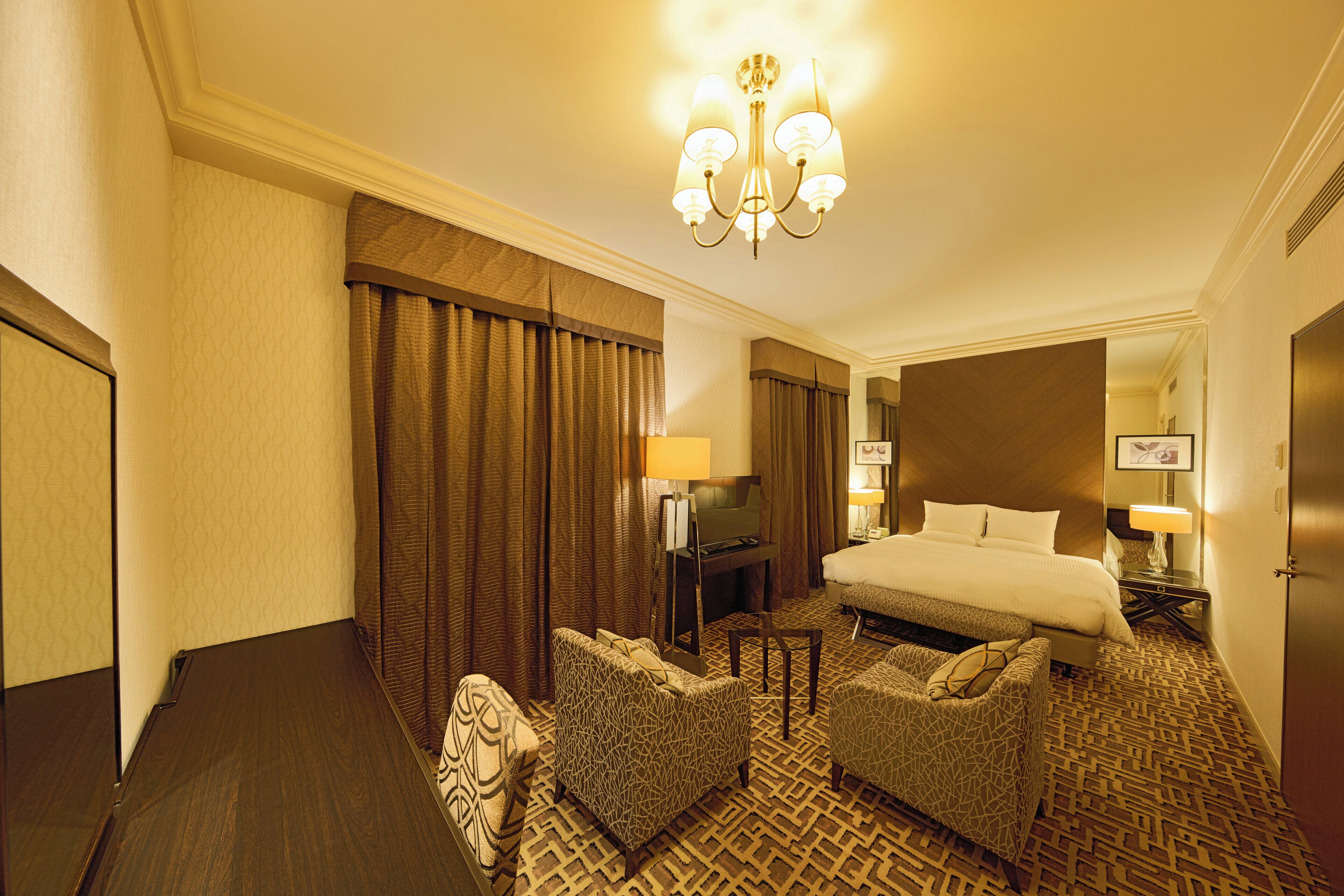 A hotel room with a large bed, two armchairs, a desk with a computer, and a chandelier.