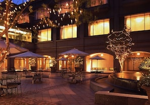Un espace extérieur aménagé en terrasse, avec des tables et des chaises, entouré d’arbres et agrémenté d’une fontaine.
