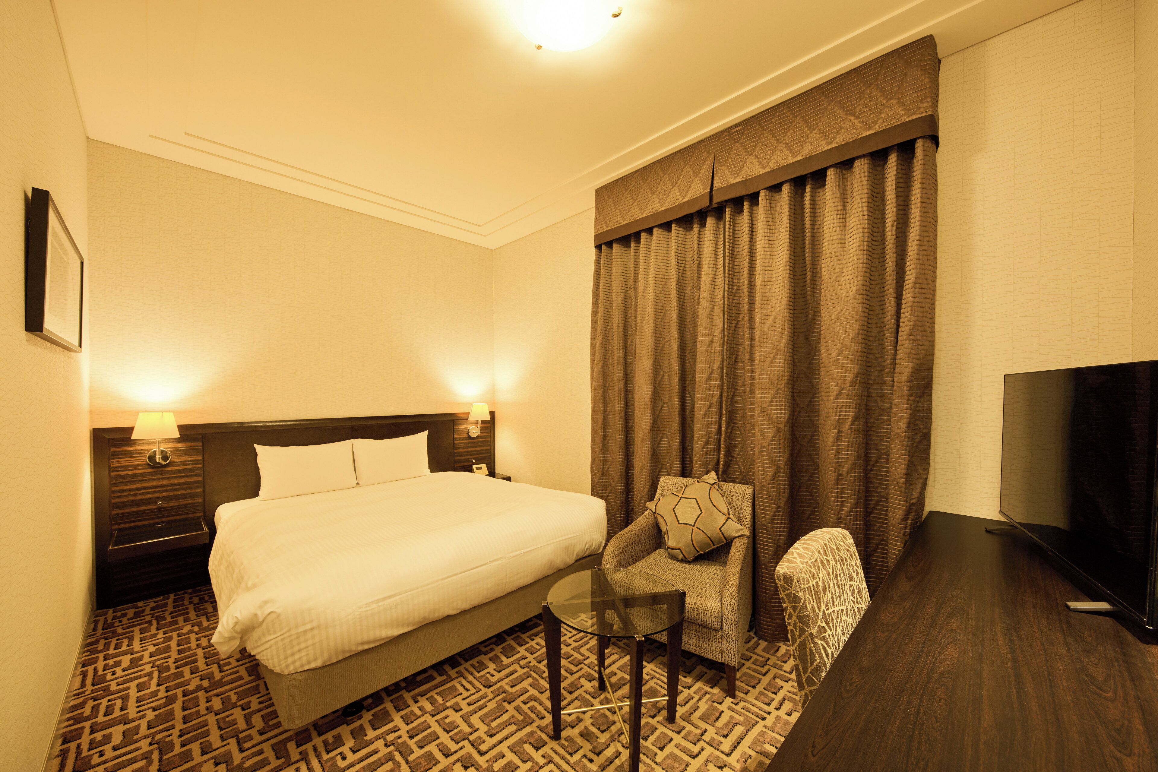 A hotel room with a bed, a chair, a small table, a TV, and a patterned carpet.