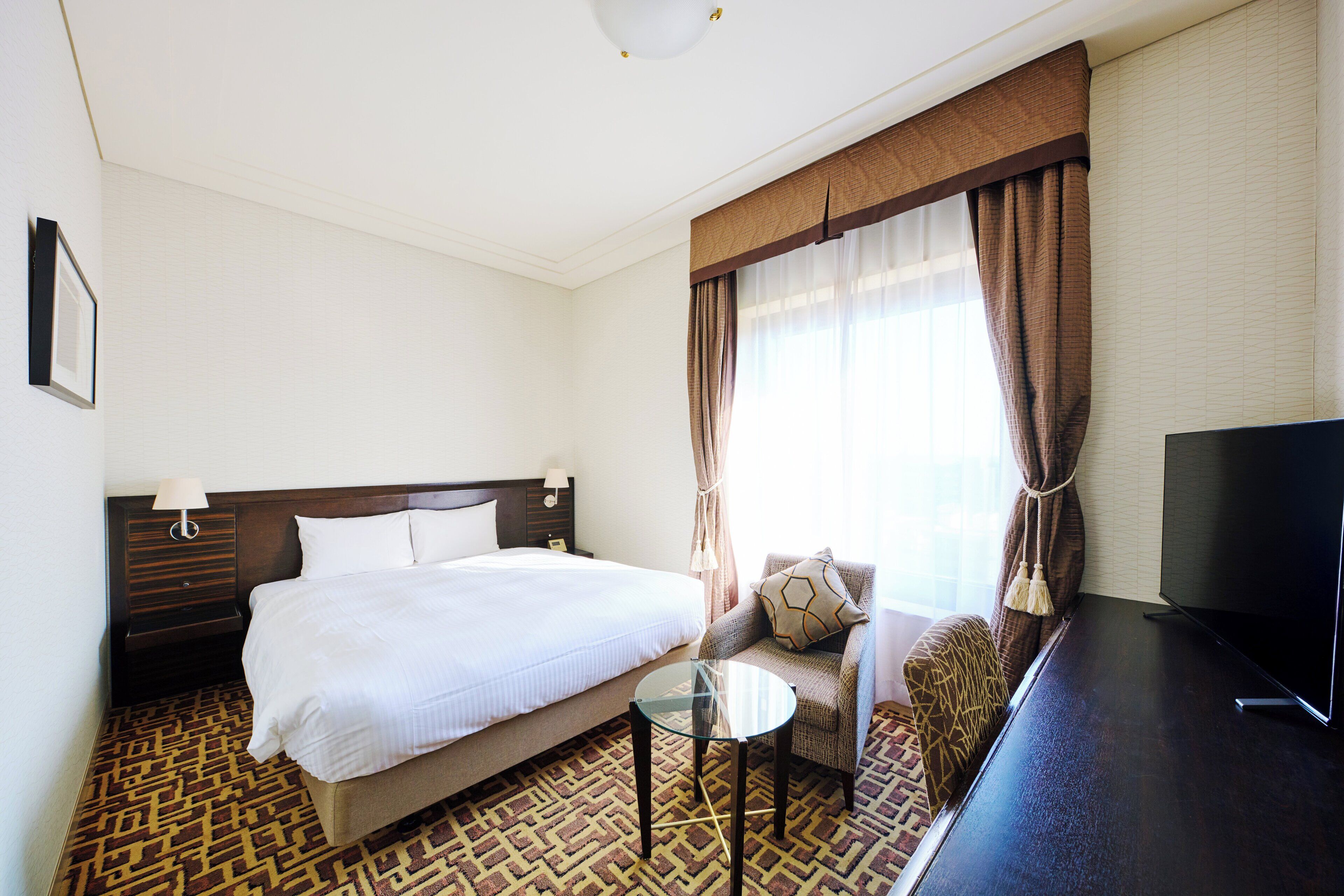 A hotel room with a large bed, a small round table, a TV, and a window with curtains.