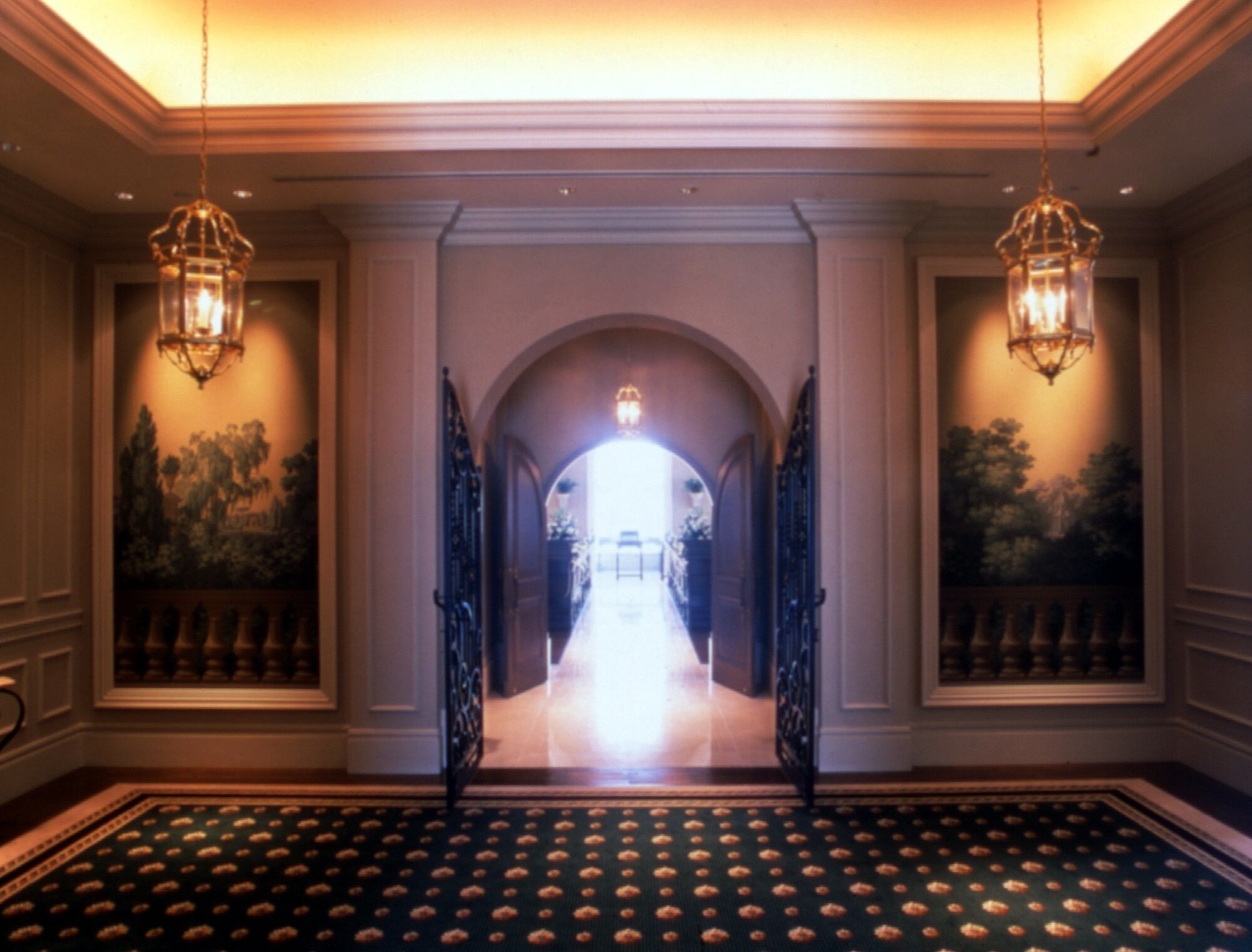 A hallway with a patterned carpet, two chandeliers, and a view into a brightly lit room.