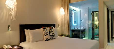 Une chambre d’hôtel avec un grand lit, un téléphone sur la table de chevet et une salle de bain fermée par des parois de verre, dotée d’un miroir.