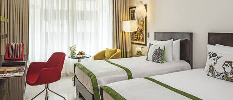 A hotel room with two beds, a desk with a chair, a lamp, and a window with curtains.