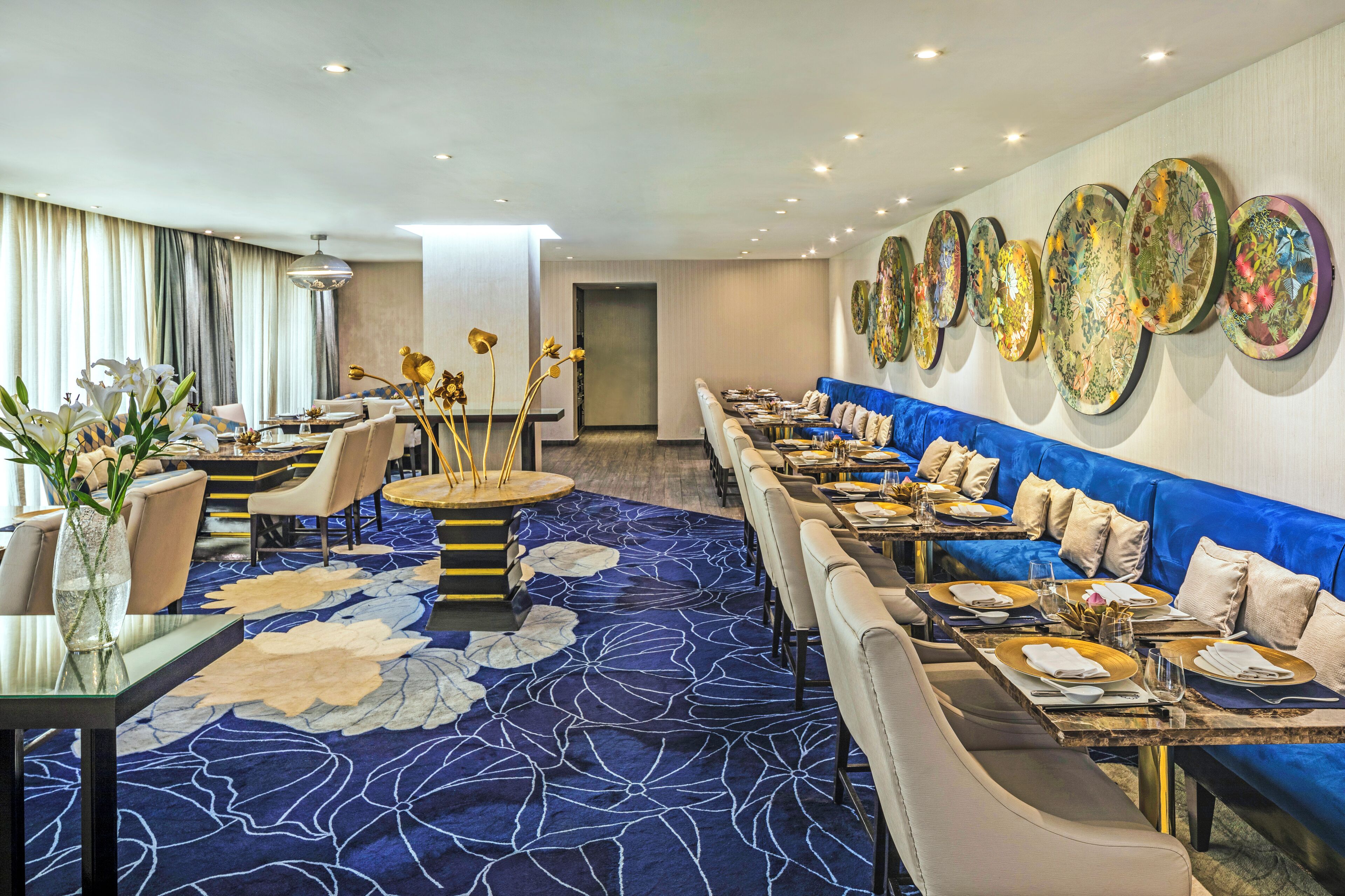 A modern hotel dining area with long tables, blue sofas, and a large floral carpet.