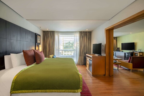 A hotel room with a large bed, a desk with a TV, and a view of the city through the window.