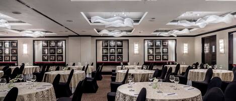 A banquet hall with round tables, black chairs, and framed pictures on the walls.