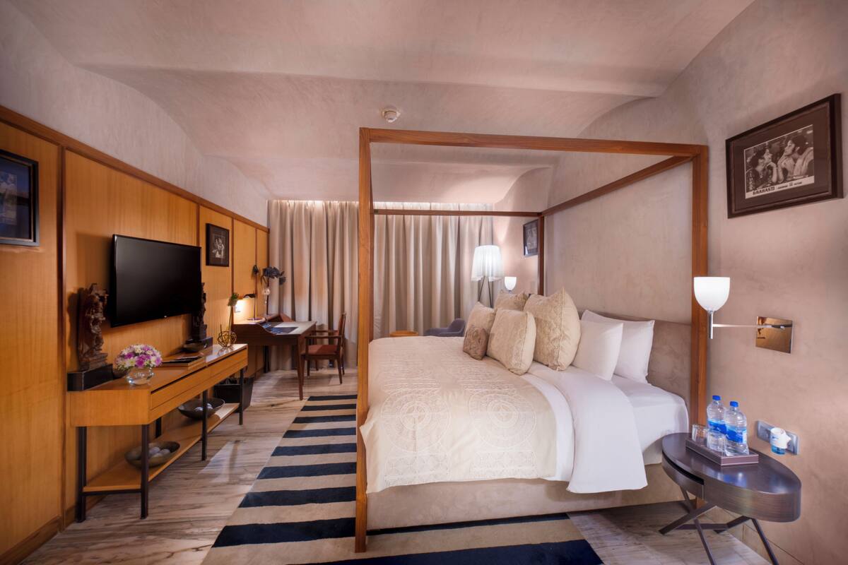 Une chambre d’hôtel comprenant un lit, un bureau avec une télévision, une chaise, une lampe et une table de chevet avec des bouteilles d’eau.