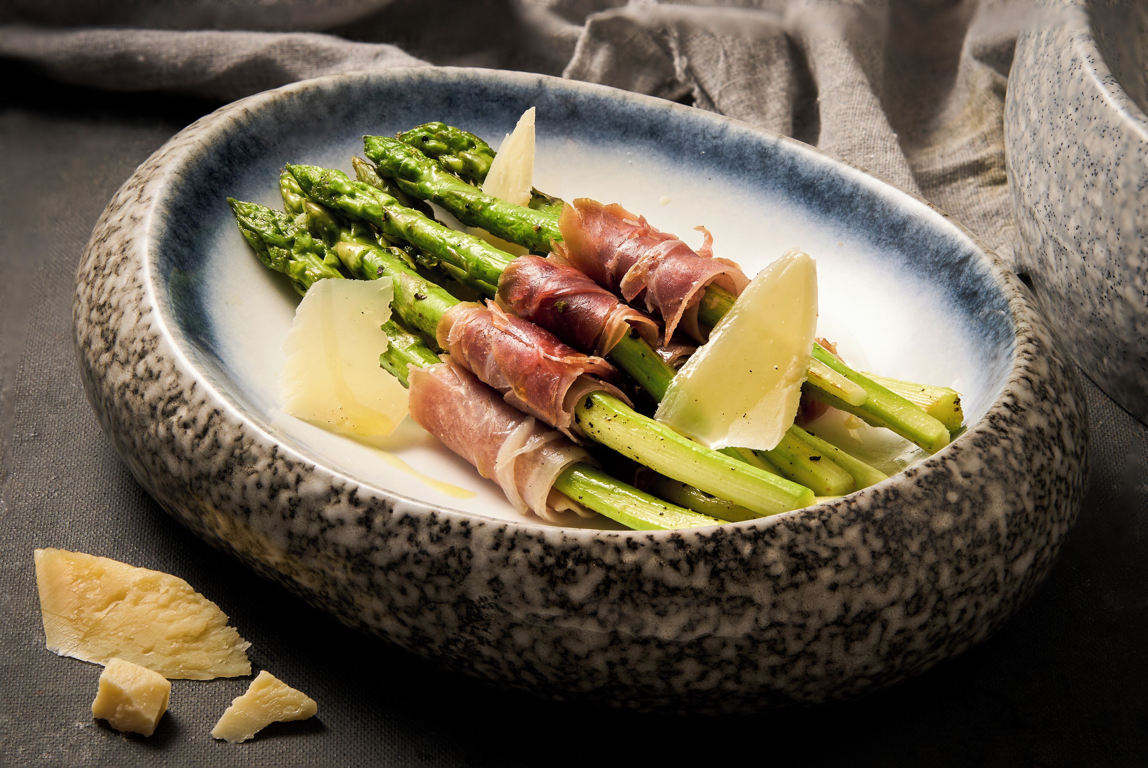 Un cuenco de espárragos envueltos en prosciutto y queso sobre una superficie oscura.