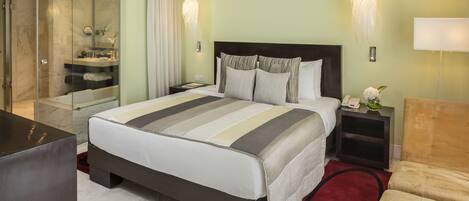 Habitación de hotel con una cama grande, baño separado con ducha de vidrio, mesita de noche con lámpara y alfombra roja.