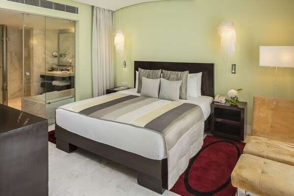 Habitación de hotel con una cama grande, baño separado con ducha de vidrio, mesita de noche con lámpara y alfombra roja.