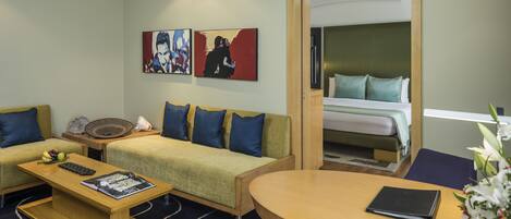 A hotel room with a sofa, a small table, a bed, and artwork on the wall.
