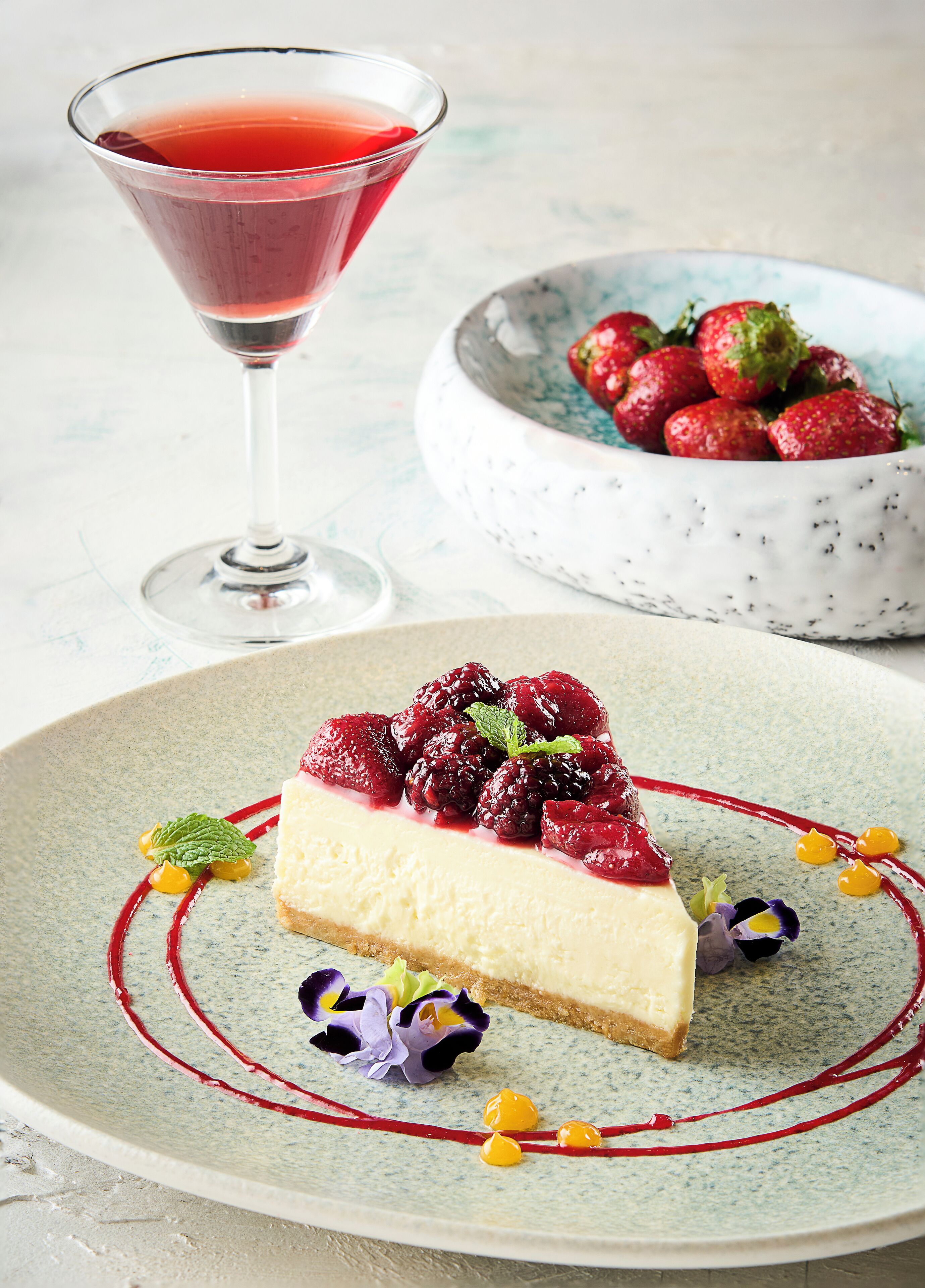 A dessert plate with a slice of cheesecake topped with berries and a red sauce, a bowl of strawberries, and a martini glass with a red drink.