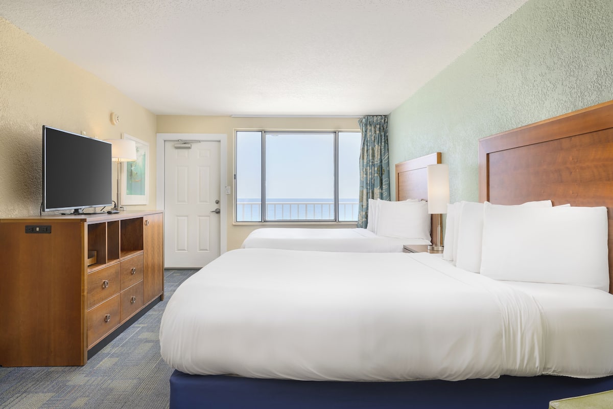 A hotel room with two beds, a TV, a wooden dresser, a window with a view of the sea, and a door.
