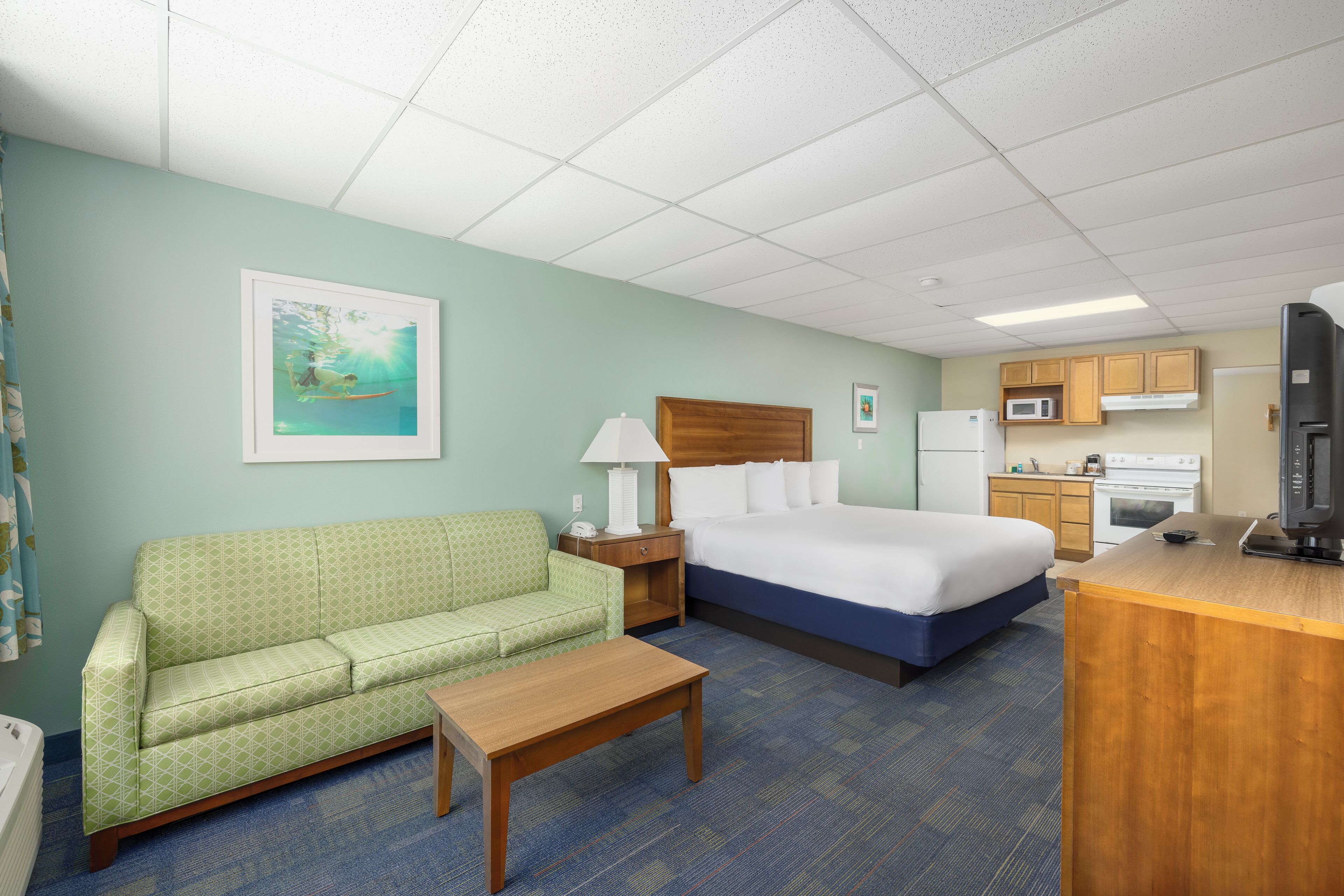 A hotel room with a green patterned sofa, a wooden nightstand, a bed with white linens, a wooden desk, and a kitchenette with a microwave and stove.