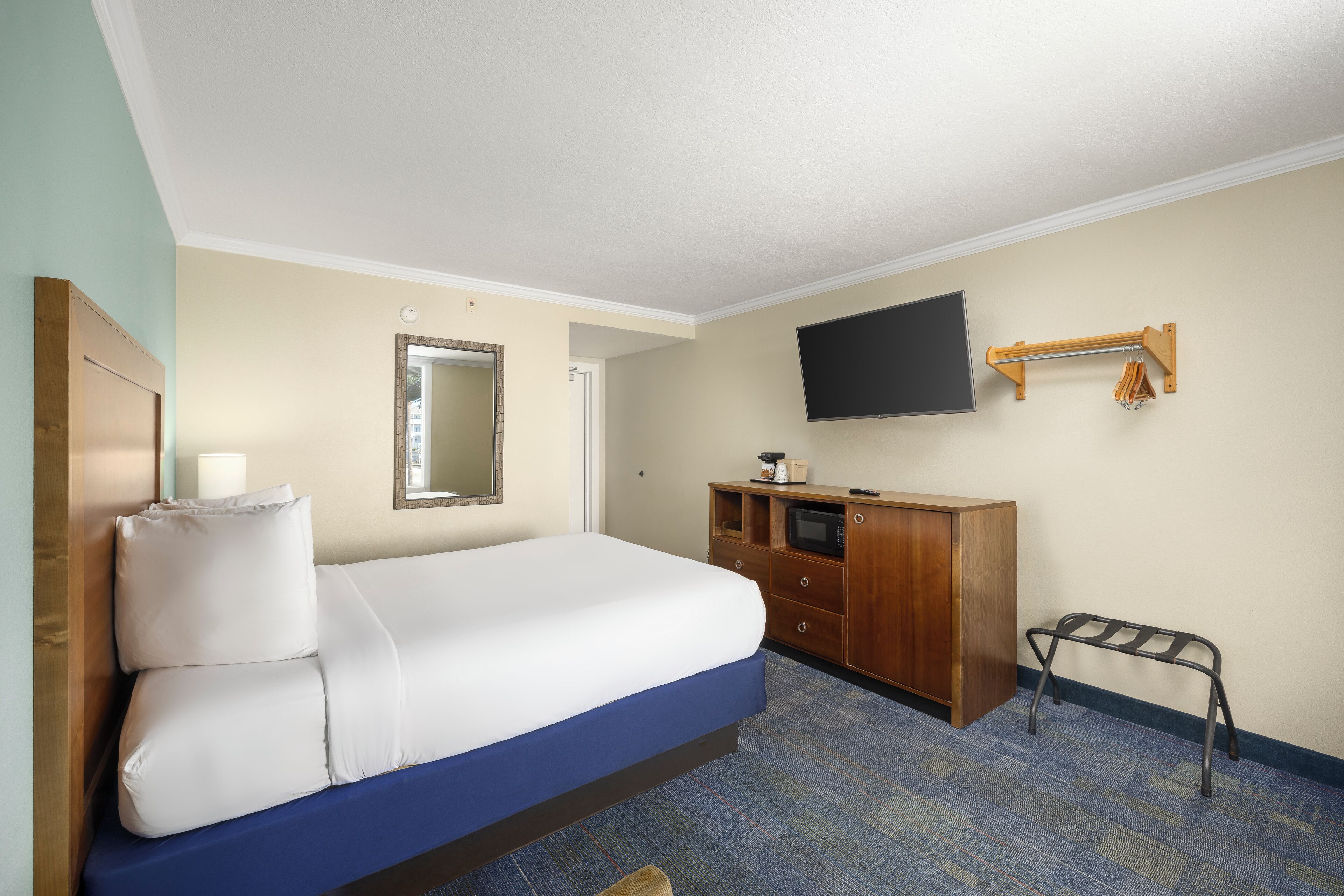Une chambre d’hôtel comprenant un lit, une commode en bois, un téléviseur fixé au mur et un petit porte-manteau.