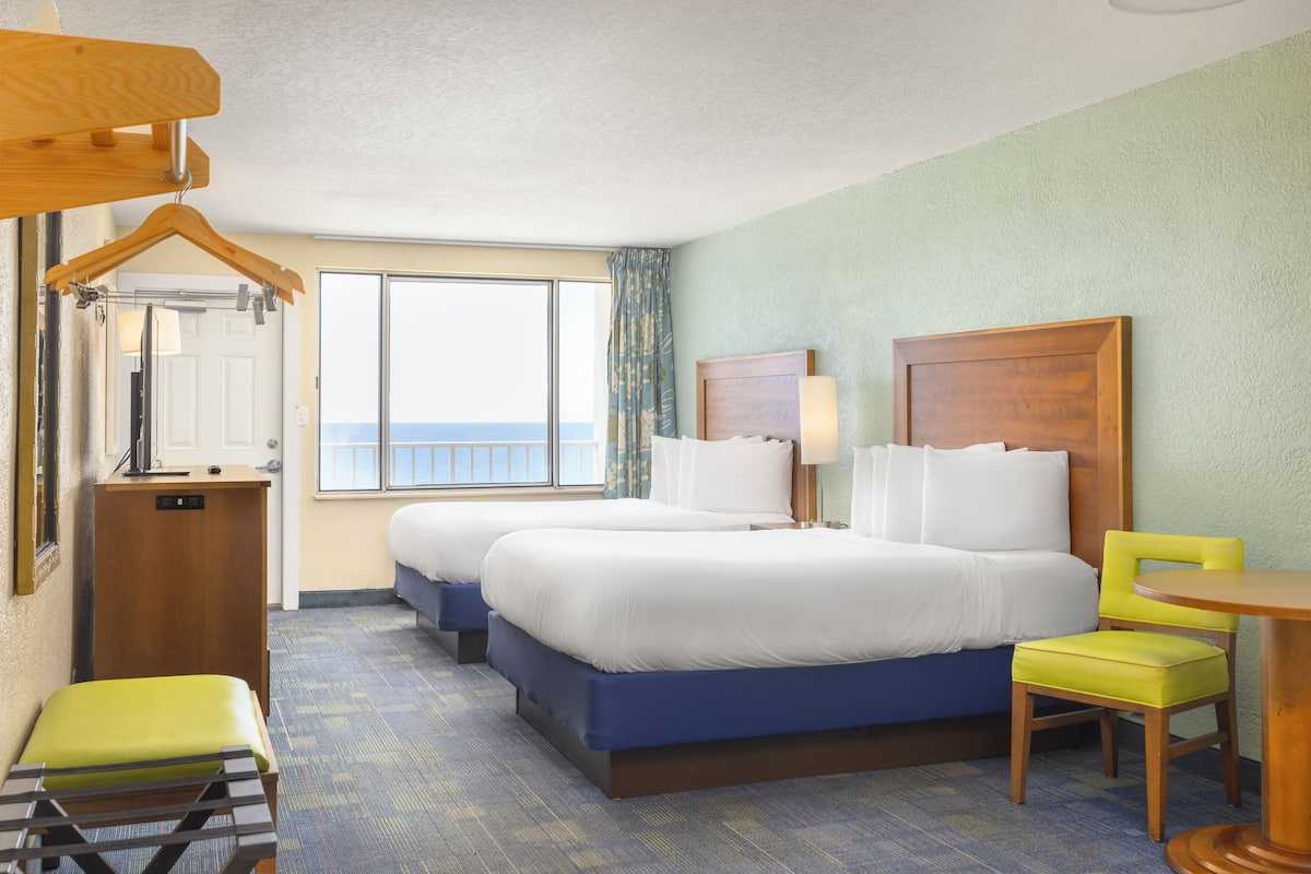 A hotel room with two beds, a desk, a chair, a small table, a hanging coat rack, and a view of the ocean.