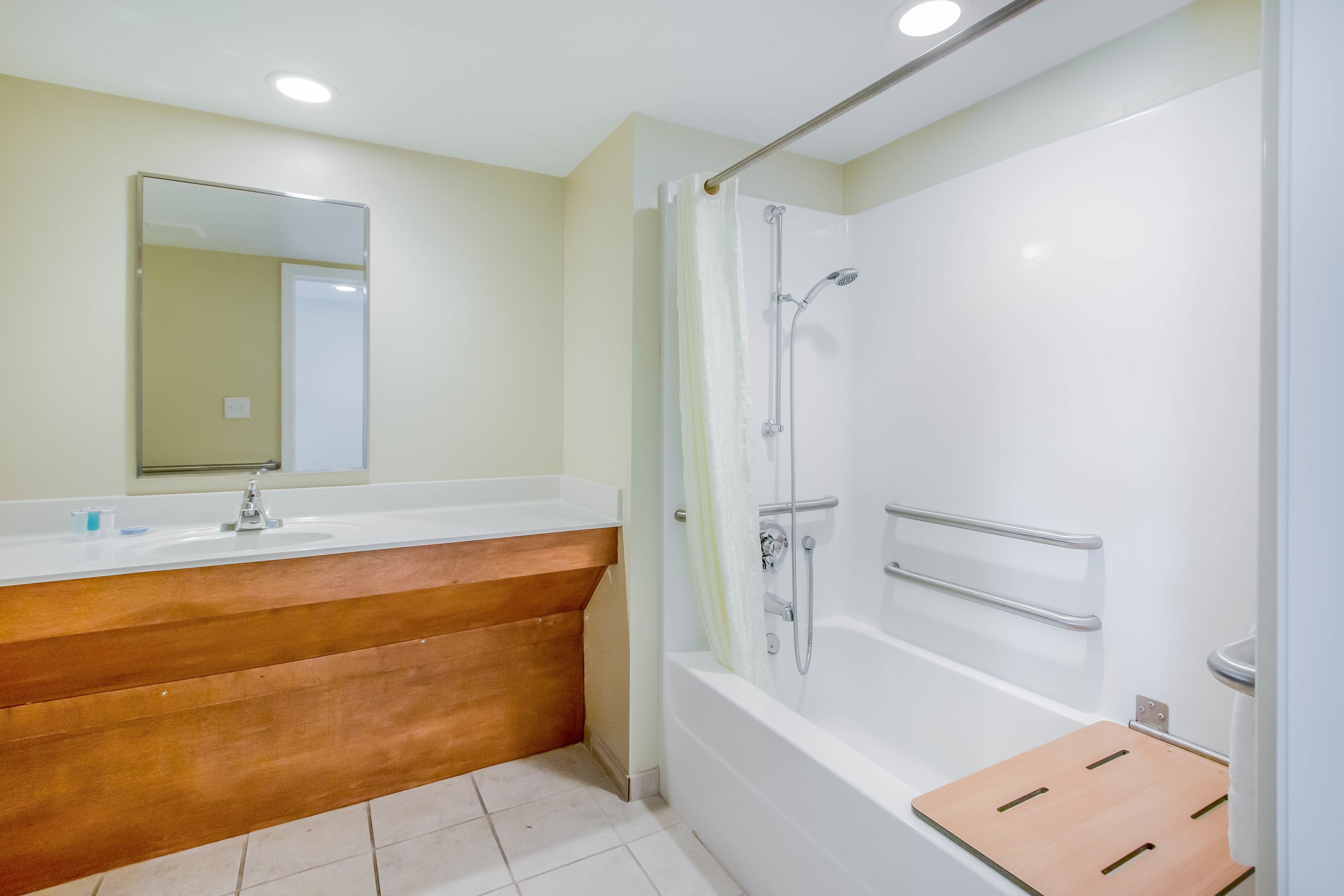 Une salle de bain avec un meuble-lavabo en bois, un évier, un miroir et une douche équipée d’une barre d’appui.