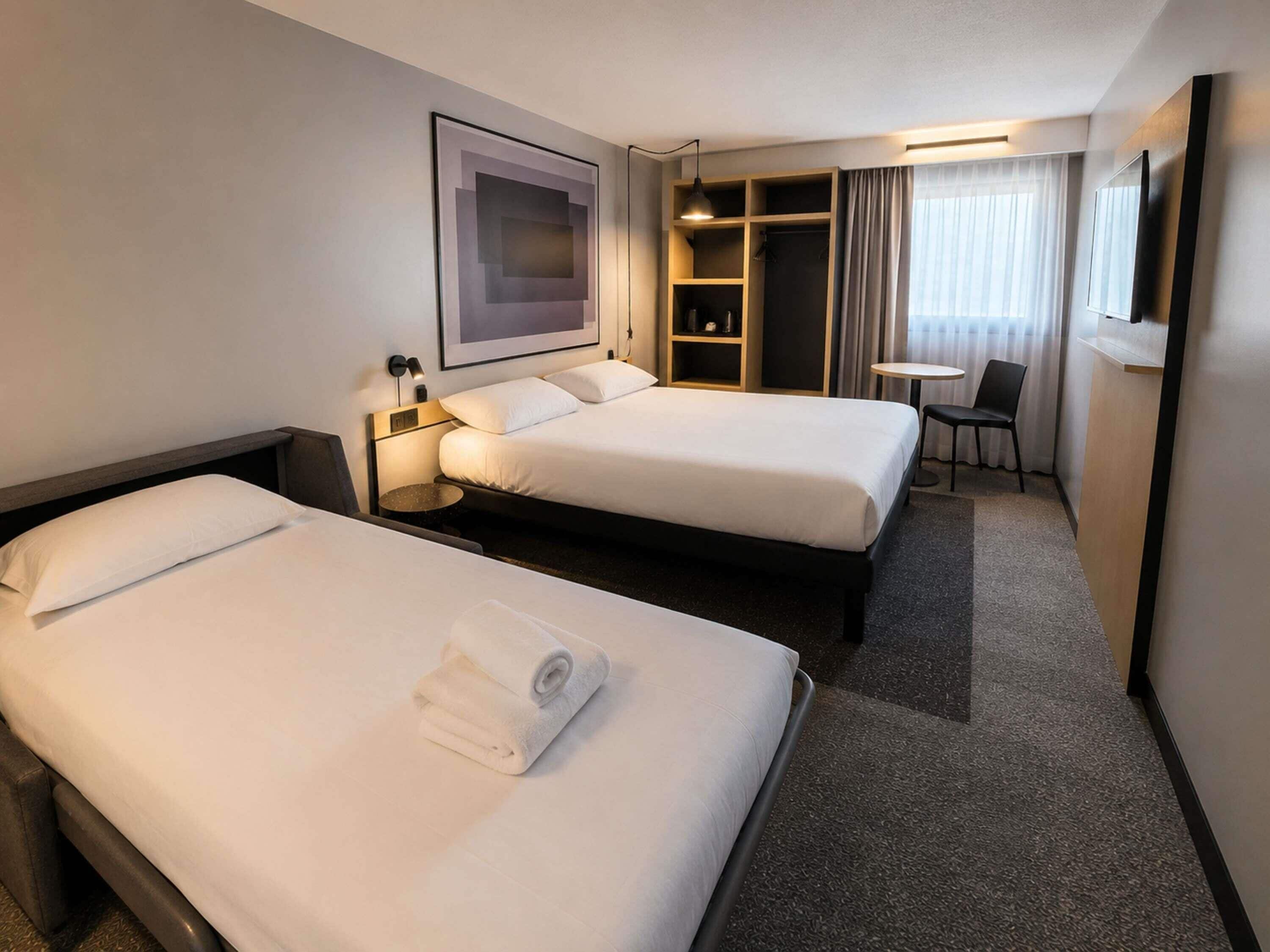 A hotel room with two beds, a desk, a chair, a small table, a wardrobe, and a framed picture on the wall.
