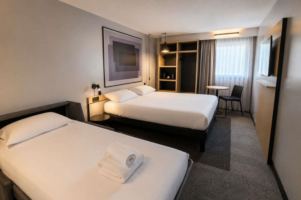 A hotel room with two beds, a desk, a chair, a small table, a wardrobe, and a framed picture on the wall.