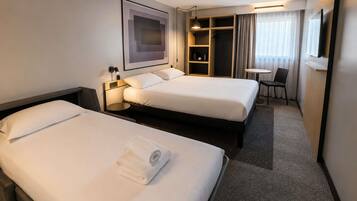 Ein Hotelzimmer mit zwei Betten, einem Schreibtisch, einem Stuhl, einem kleinen Tisch, einem Kleiderschrank und einem gerahmten Bild an der Wand.