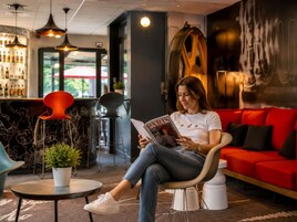 A woman reading a magazine in a modern lounge area with a red couch, a bar with bottles, and a large abstract wall art piece.