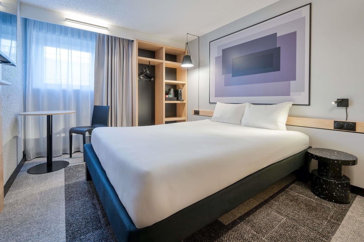 A modern hotel room with a large bed, a desk, a chair, a wardrobe, and a wall-mounted artwork.