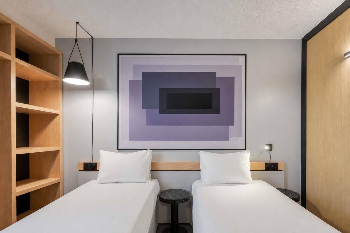 A hotel room with two beds, a wooden wardrobe, a round table, and a modern art piece on the wall.