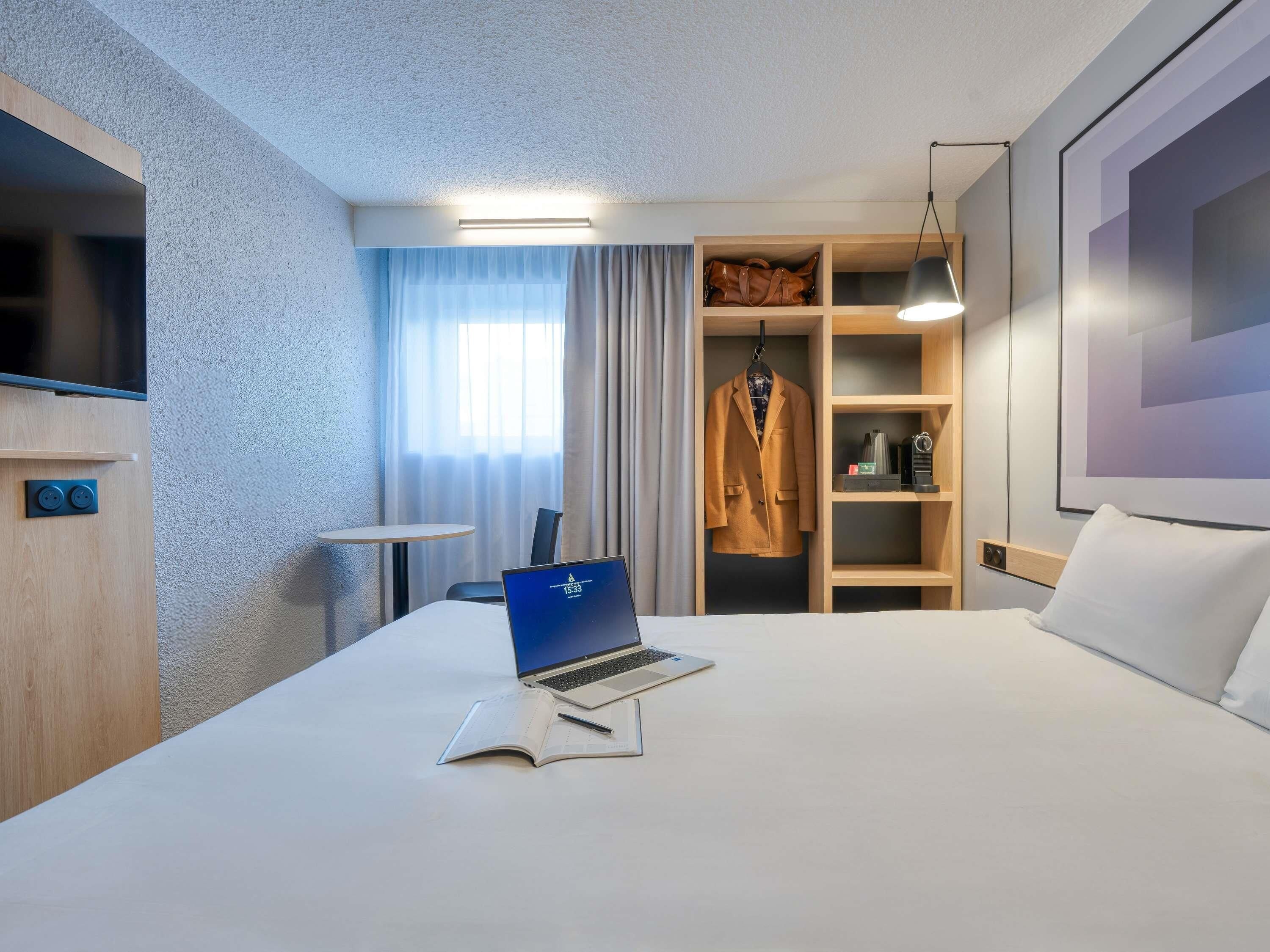 Habitación de hotel con cama, escritorio con computadora portátil, televisor, armario y una pequeña mesa.