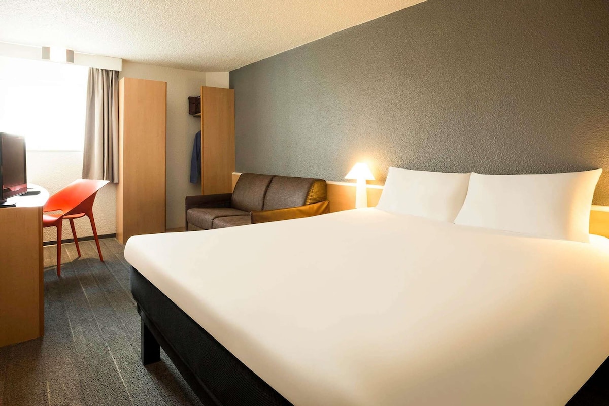 Una habitación de hotel con cama, escritorio con silla, televisor y ventana con cortinas.