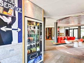 A modern lobby with a vending machine, red seating area, and a large poster. - Hôtel ibis Orly Chevilly Tram 7 (Chevilly-Larue)