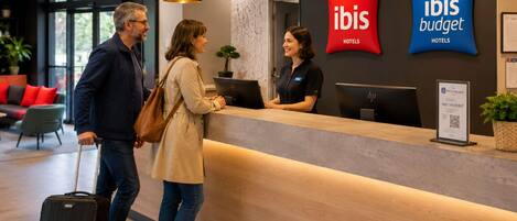 A hotel reception desk with 'ibis Hotels' branding, a receptionist, and guests.