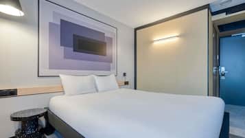 Habitación de hotel con una cama, una mesita auxiliar, una lámpara adosada a la pared, una obra de arte abstracta enmarcada y una puerta que da a otra habitación.
