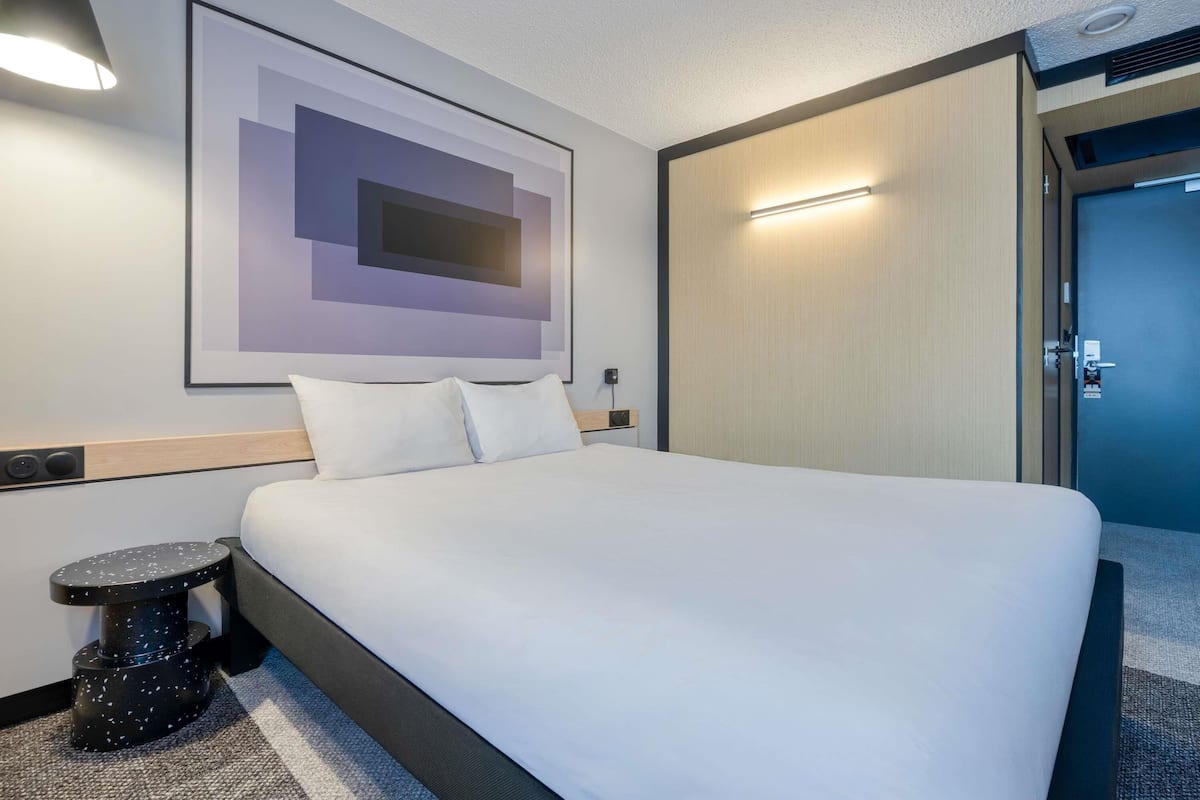 A modern hotel room with a large bed, a side table, a wall-mounted light, and a door to the right.