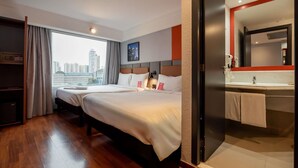 Habitación de hotel con dos camas, un ventanal grande con vistas a la ciudad, baño con espejo y lavamanos, y suelo de madera.