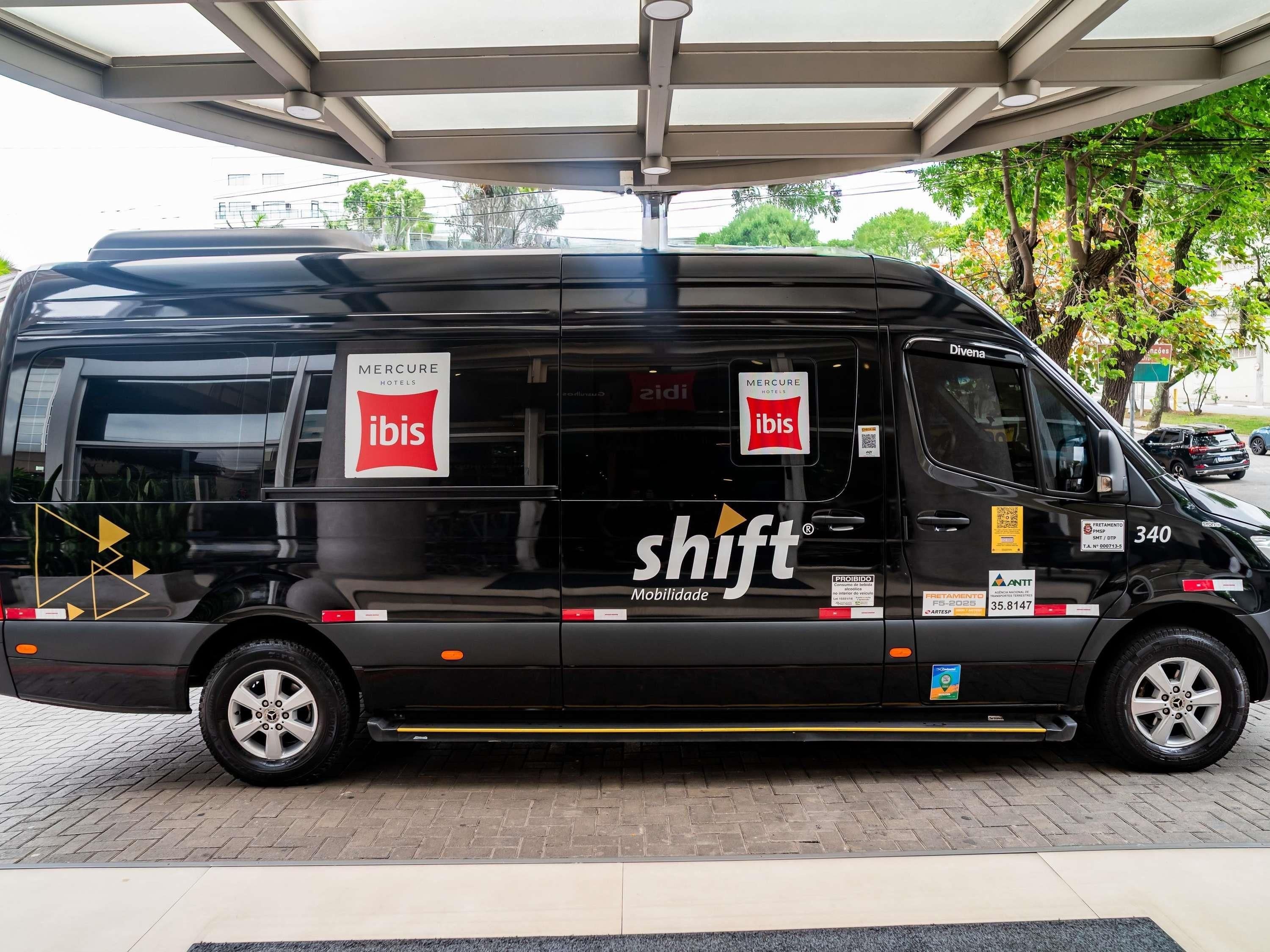 Un fourgon noir arborant les logos « ibis » et « shift Mobilidade », stationné sous un abri.