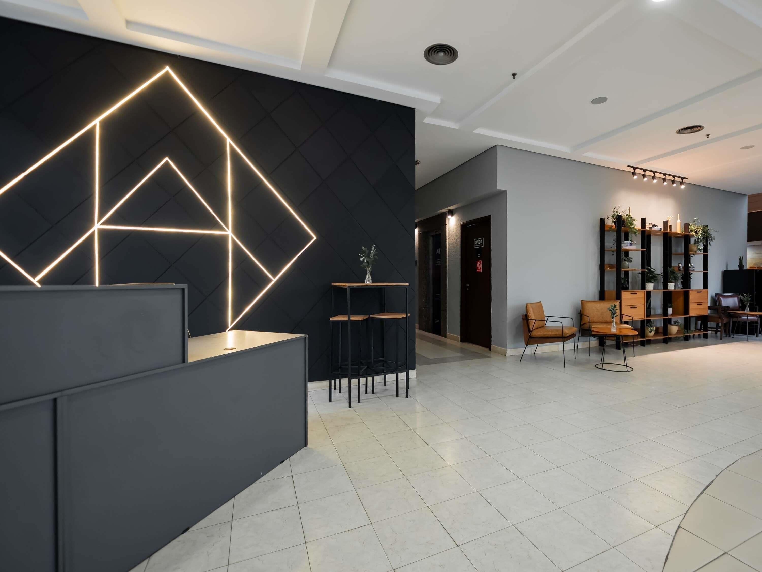 Uma área de recepção moderna com uma instalação de iluminação geométrica, um balcão de recepção e um espaço de estar com cadeiras e uma mesa.