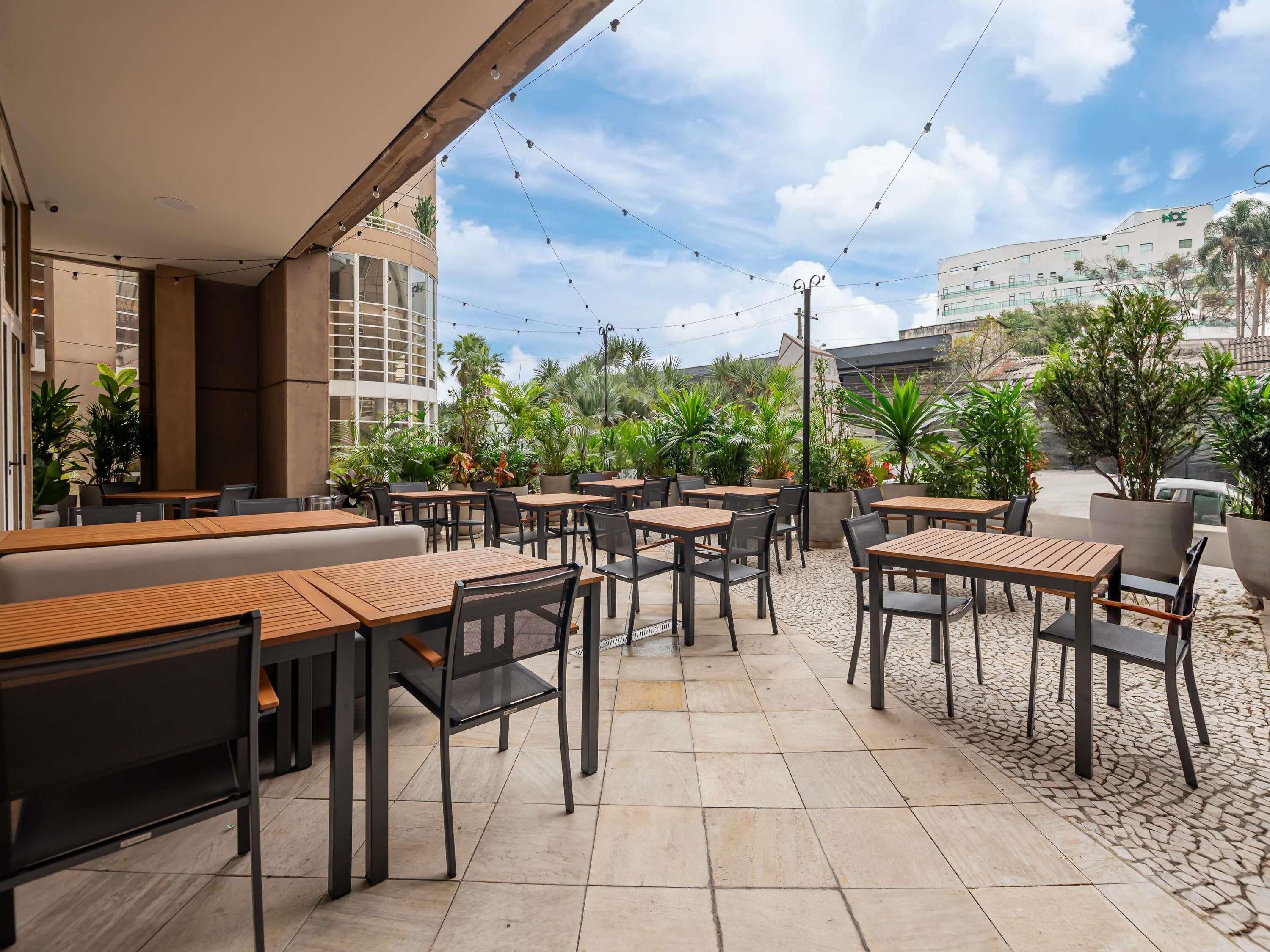 An outdoor cafe area with multiple tables and chairs, surrounded by potted plants and a view of a building in the background.
