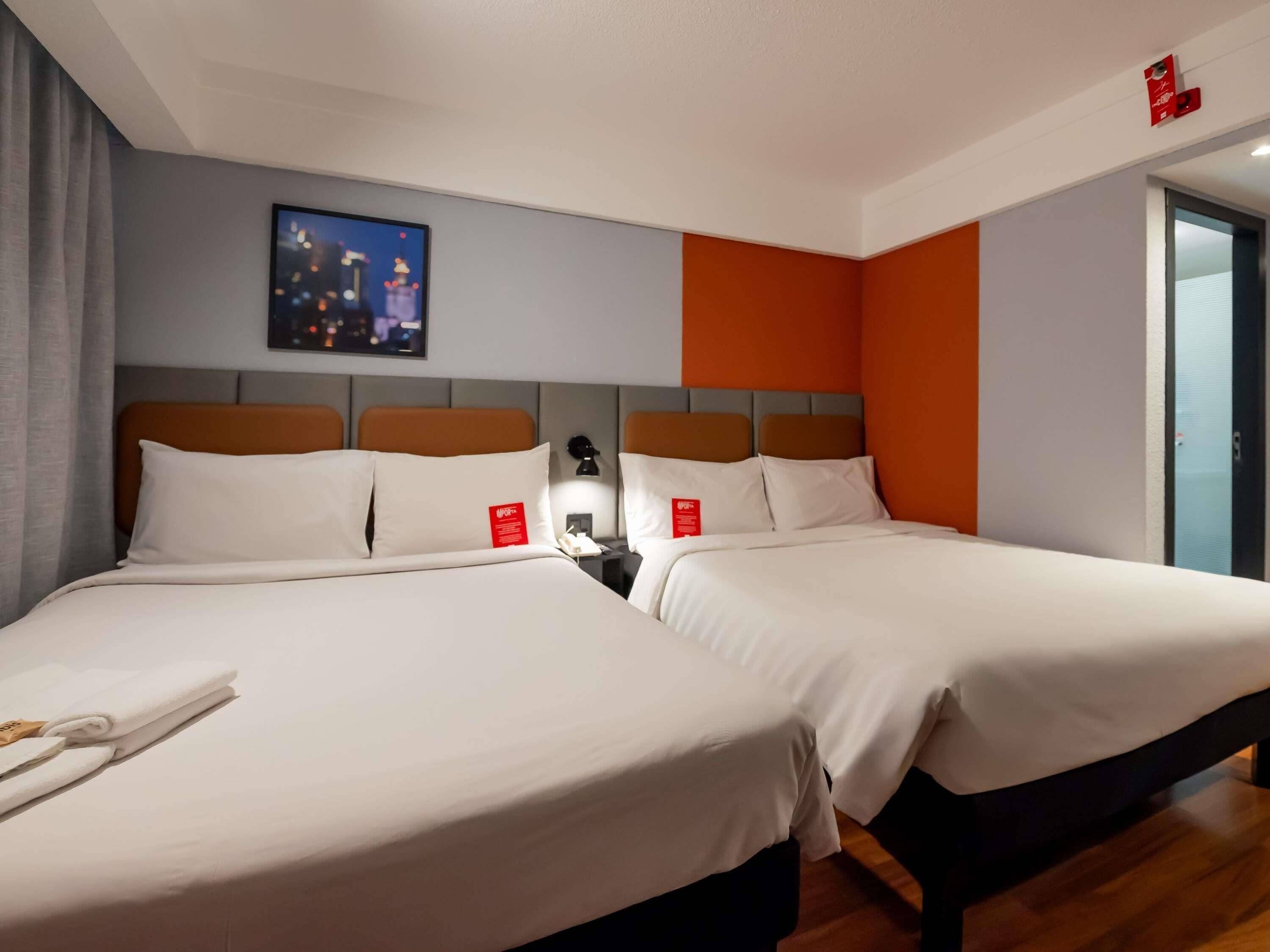 Ein Hotelzimmer mit zwei Betten, einem Nachttisch mit Lampe, einem Stadtbild als Gemälde und einer Tür mit rotem Notlicht.