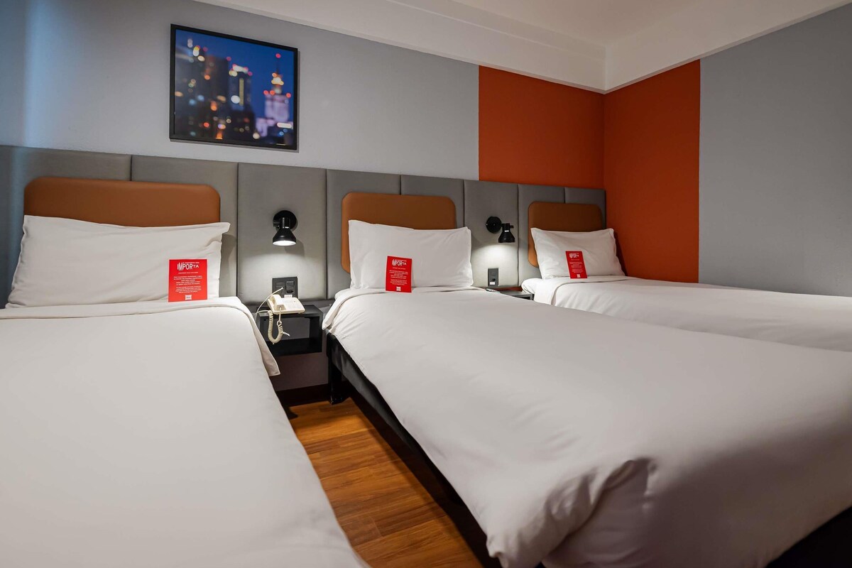 A hotel room with two beds, each with white bedding and a red sign. There is a nightstand with a phone and a lamp between the beds. A painting of a cityscape is on the wall above the beds.