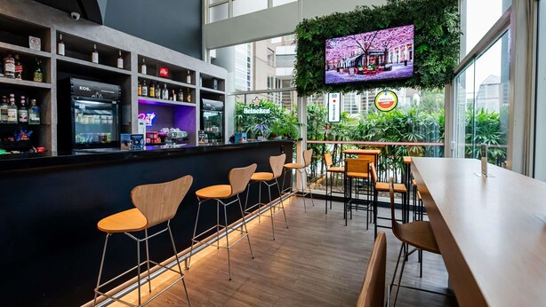 Zona de bar moderna con una pantalla grande mostrando un paisaje urbano, taburetes de madera y una variedad de botellas de licor.