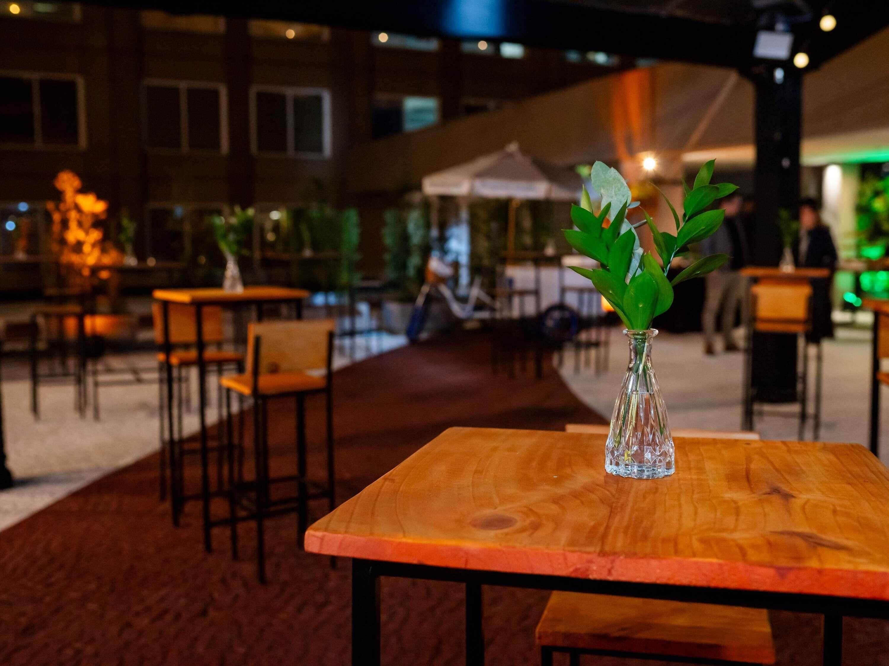 Una mesa de madera con un florero de vidrio transparente que contiene hojas verdes, ubicada en un espacio interior tenuemente iluminado con sillas y mesas dispuestas alrededor.