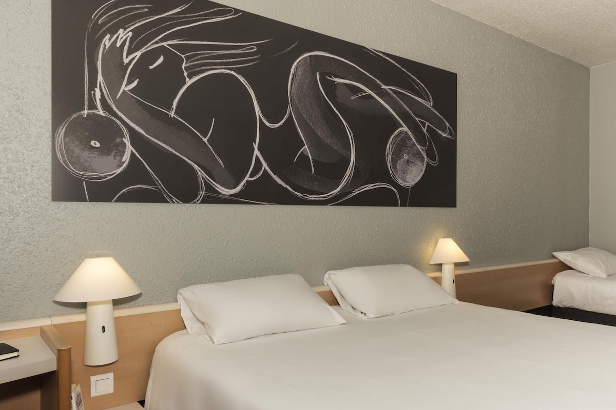 Quarto de hotel com cama, duas luminárias de cabeceira, uma obra de arte na parede e uma mesa de cabeceira com abajur.