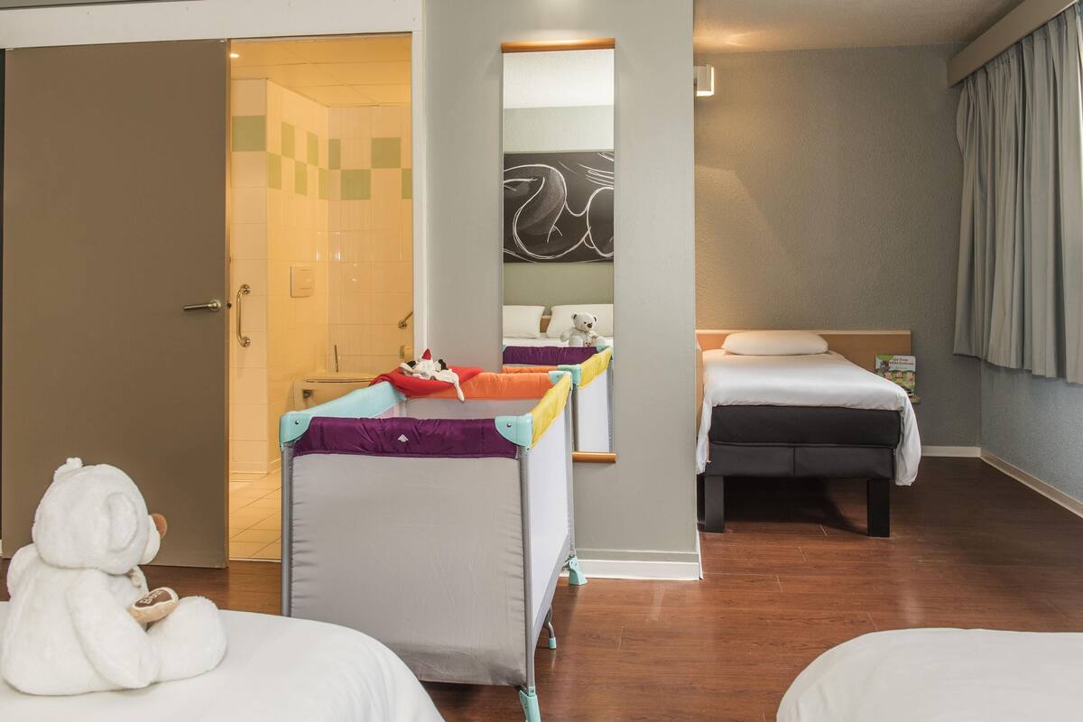 Quarto de hotel com uma cama, uma secretária pequena, uma cadeira, banheiro com chuveiro e um espelho.