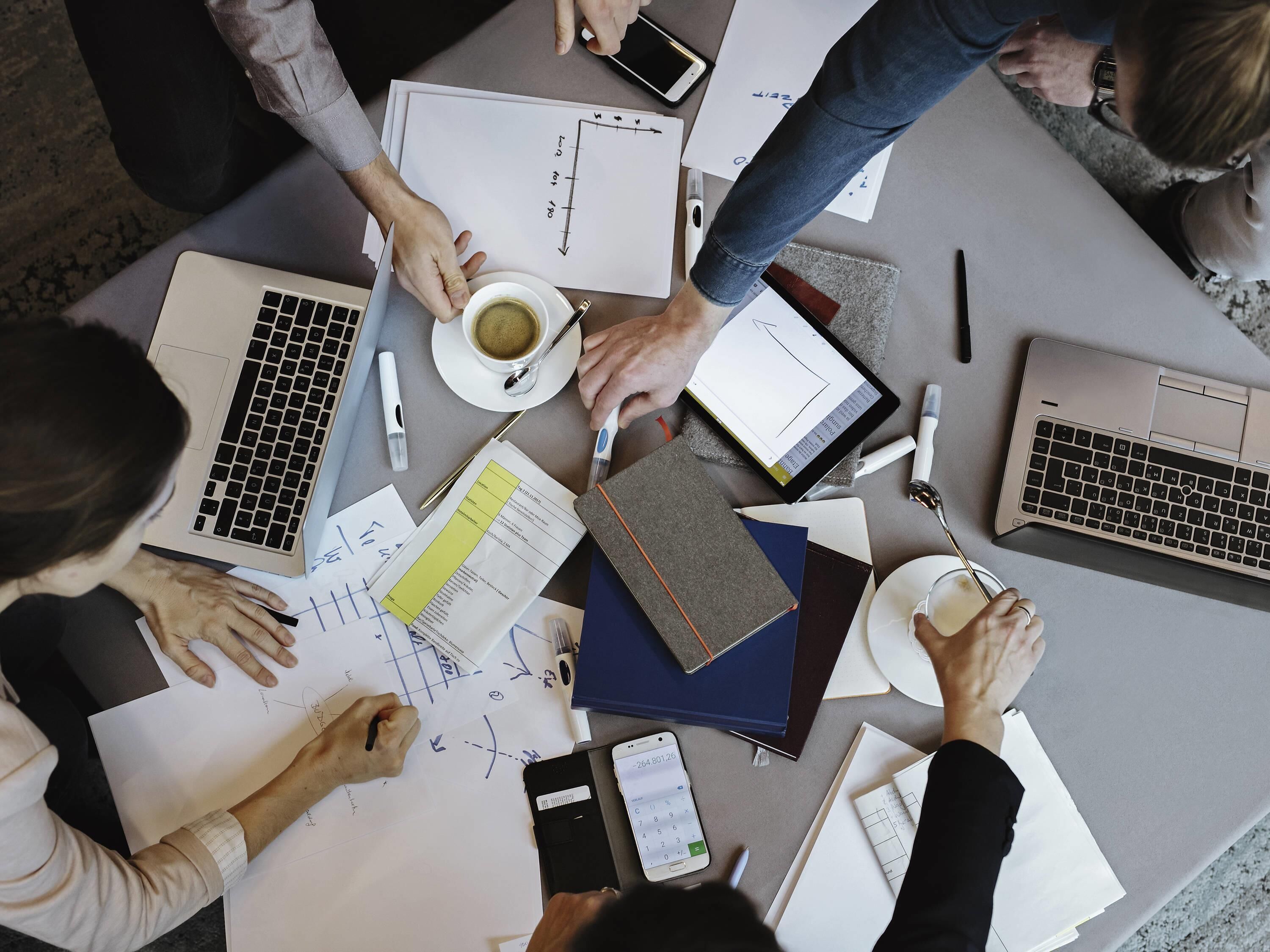 Un groupe de personnes est réuni autour d’une table, devant des ordinateurs portables, des documents et un smartphone.
