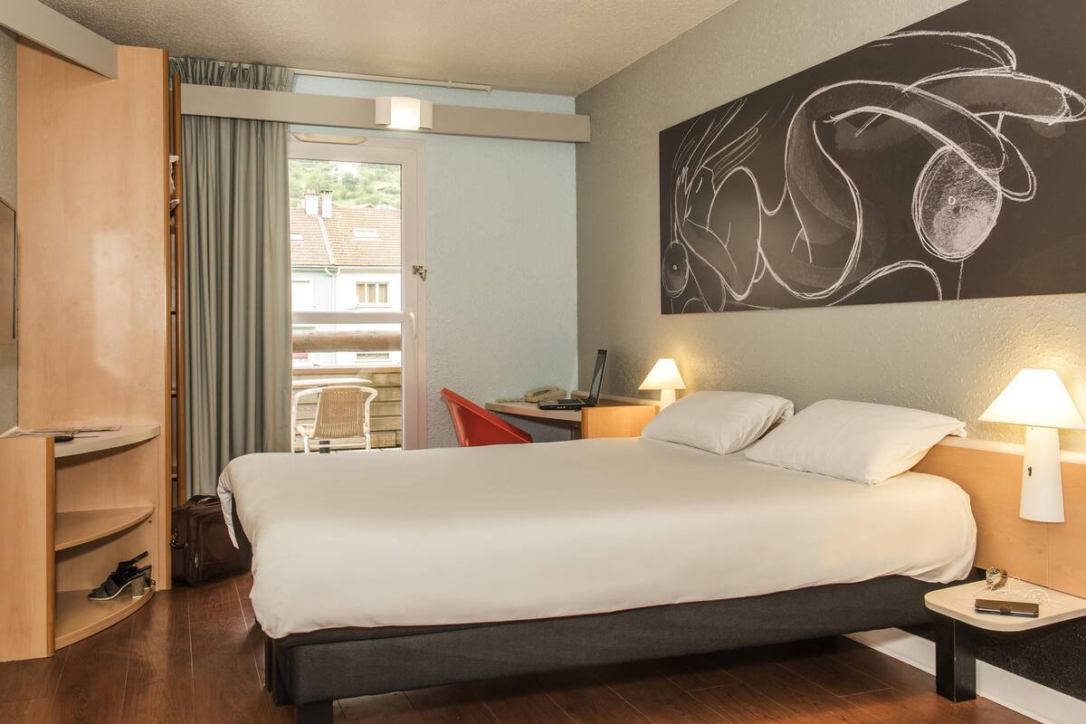 Quarto de hotel com cama, escrivaninha com luminária, cadeira, guarda-roupa e uma grande pintura abstrata na parede.